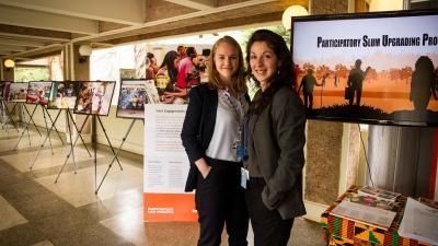 Two women smiling in a hallway next to an exhibition on "Participatory Slum Upgrading Project" with photos.