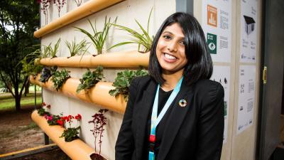 A smiling woman stands next to a vertical garden made of bamboo with plants, promoting sustainability.