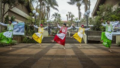 Five men in traditional colorful outfits perform a dance outdoors on a paved area with buildings in the background.