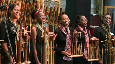 Five women smiling and playing traditional wooden instruments on stage.