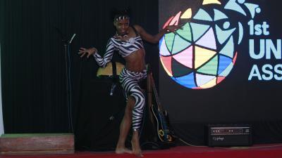 A performer in a black and white striped outfit poses on stage in front of a screen with a colorful globe logo.