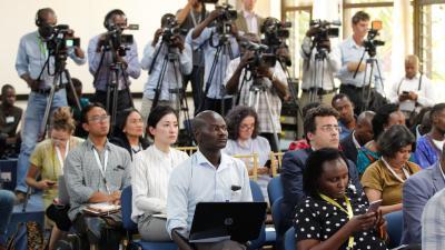 A diverse group of journalists and media personnel attend a press conference, with cameras set up.