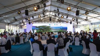 A large reception tent filled with people seated at tables, facing a stage with a large screen.