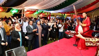 A musician in a red outfit plays an instrument on stage while people applaud at a festive reception.