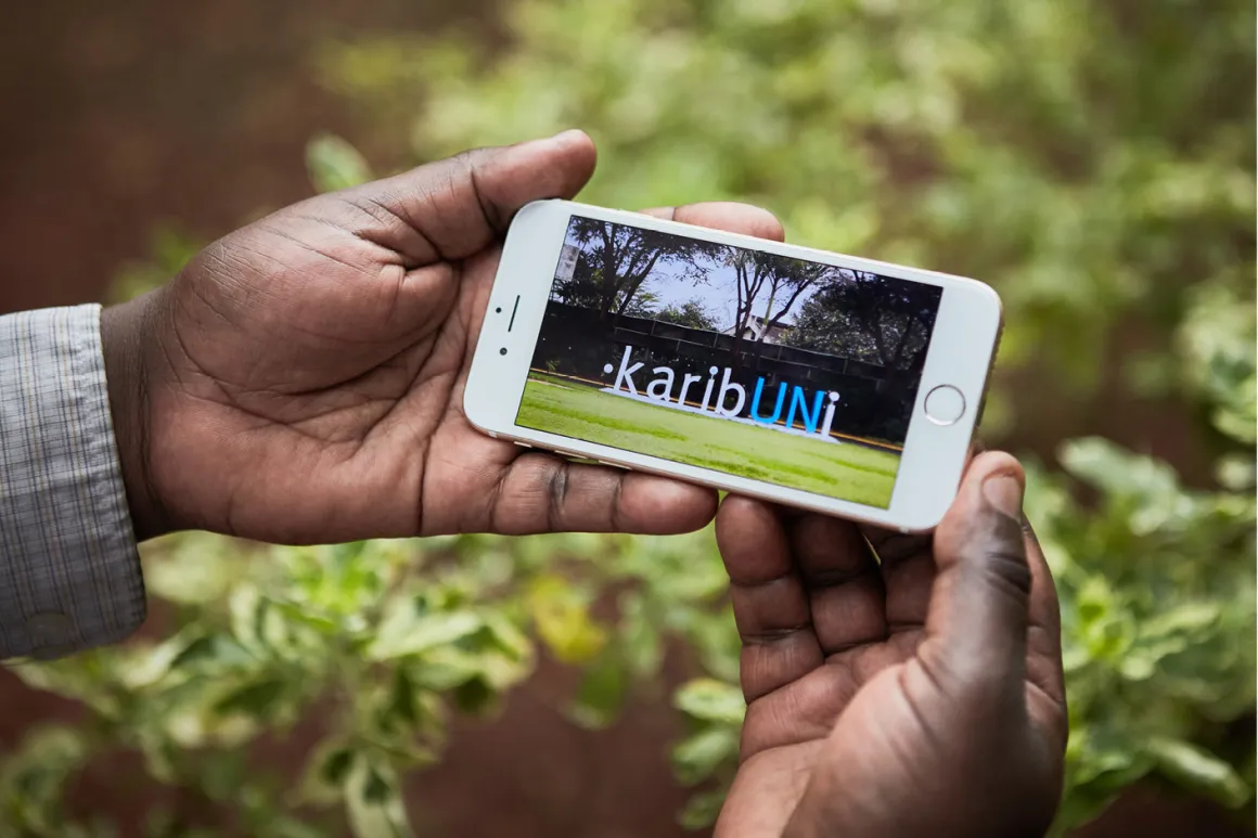 A pair of hands holding a mobile phone displaying a photo of the “karibUNi” sign installation that welcomes visitors to the UNON main lobby. The [sign?] was designed by the UNON Division of Conference Services Publishing Services Section