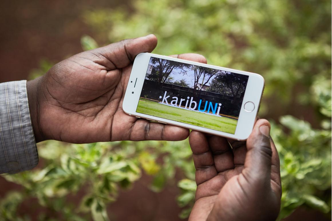 Paire de mains tenant un téléphone portable affichant une photographie du message « karibUNi » qui souhaite la bienvenue aux visiteur(se)s dans la zone centrale d’accueil de l’ONUN. L’installation a été conçue par la Section des services de publication de la Division des services de conférence de l’ONUN 